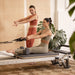 Aluminium Reformer Mentor With A Woman Performing The Exercise Using The Resistance Band And Instructor With Potted Green Plants In The Background