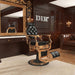 DIR Princeton Gold Barber Chair Black Variant Front Angled View With Big Decorative Mirror A Patent Sofa And A Brand Name In The Background