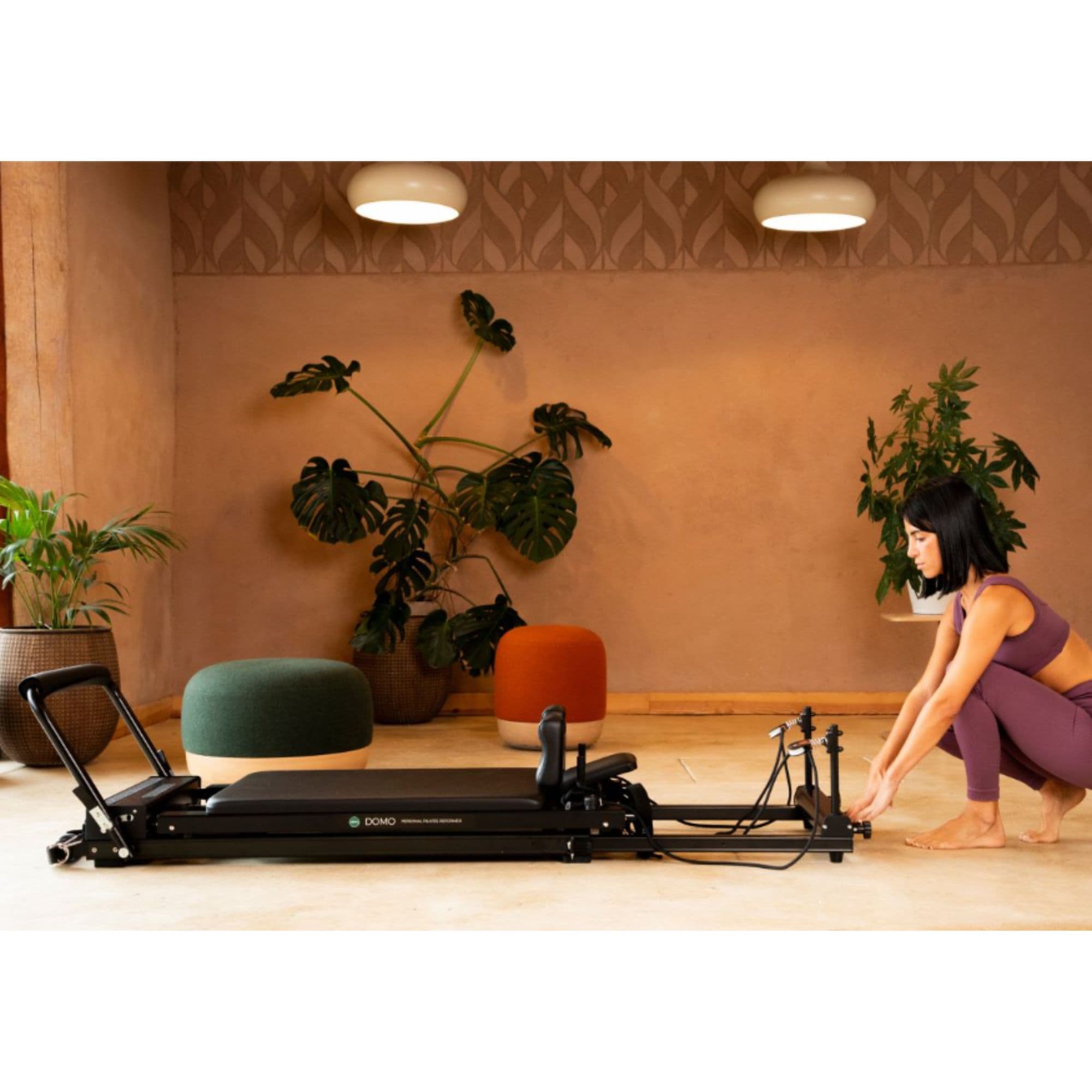 Elina Domo Home Reformer Side View Of A Woman Adjusting Ropes With Potted Green Plants In The Background
