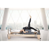 Thumbnail for Elina Pilates Elite Wood Reformer with Tower With A Woman Lying Down While Raising Her Right Legs And Huge Buildings In The Background