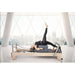 Elina Pilates Elite Wood Reformer with Tower With A Woman Lying Down While Raising Her Right Legs And Huge Buildings In The Background