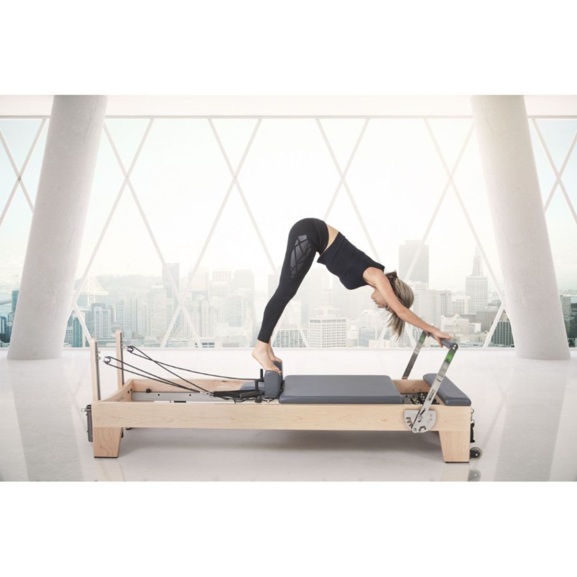 Elina Pilates Elite Wood Reformer with Tower With A Woman Performing A Front Bend Exercise And Huge Buildings In The Background