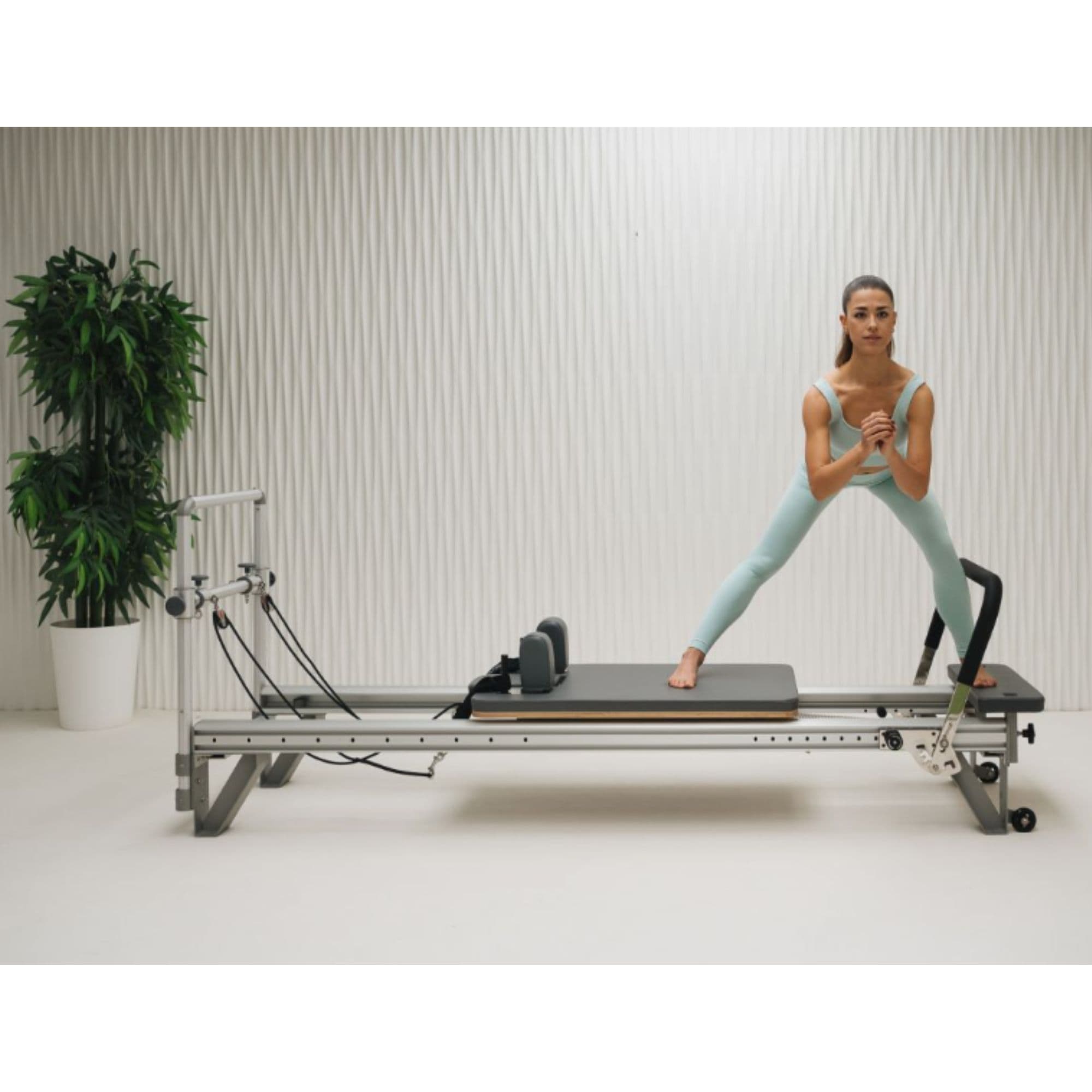 Elina Pilates Mentor Aluminium Reformer With A Woman Performing A Standing Pilates Exercise With A Potted Green Plant In The Background