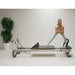 Elina Pilates Mentor Aluminium Reformer With A Woman Performing A Standing Pilates Exercise With A Potted Green Plant In The Background