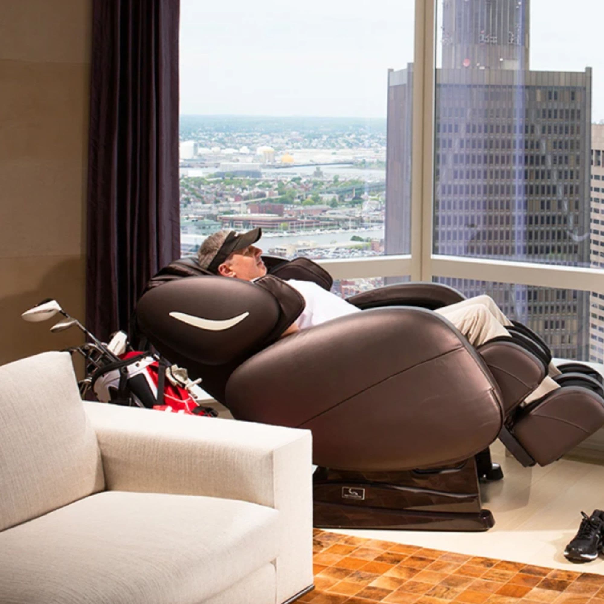 Infinity Smart Chair X3 Massage Chair Brown Variant Side View With A Couch And A Man Relaxing In The Massage Chair On Huge Building Background