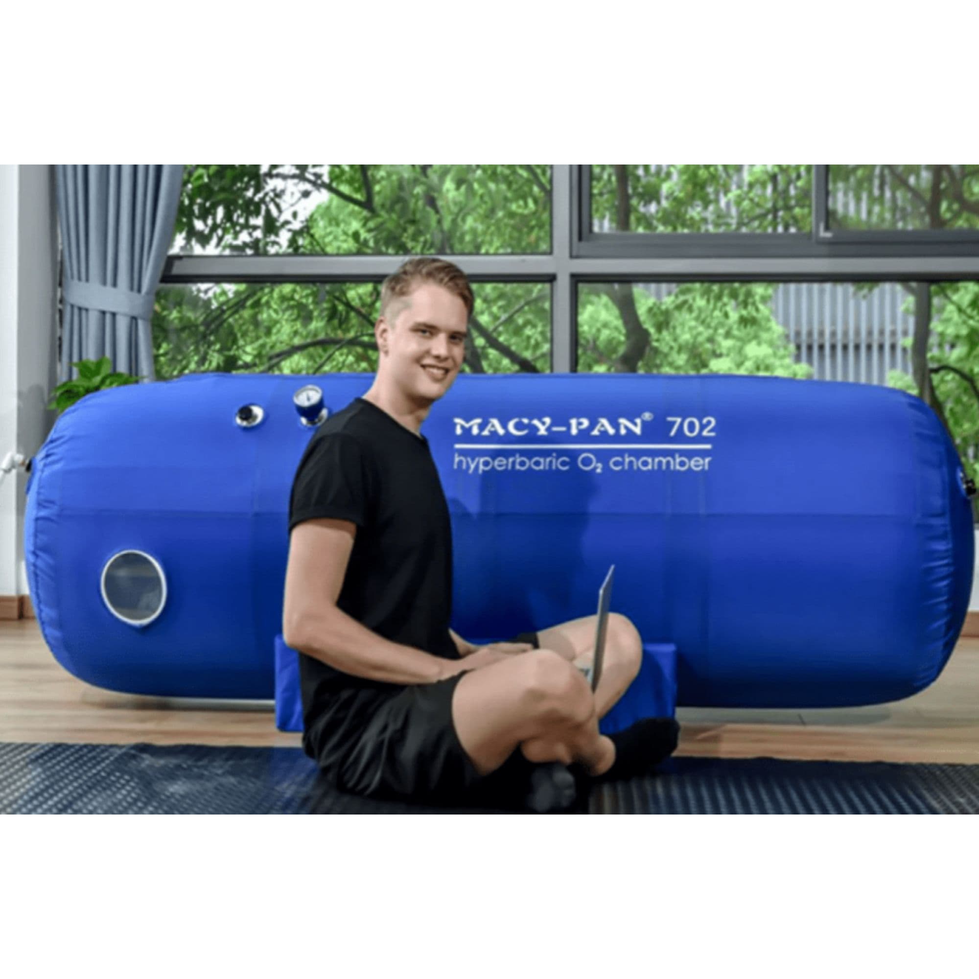 Macy-Pan ST702 Soft-Shell Hyperbaric Chamber With A Boy Sitting In The Floor While Using Laptop By Large Glass Windows With Greenery Views
