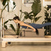 Reformer Master Instructor Side View Of A Woman Performing A Pilates Exercise On A Reformer With Tropical Green Plants In The Background